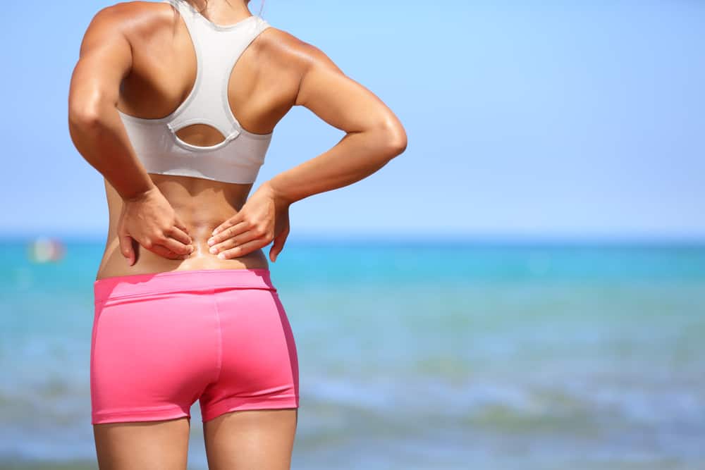 Athletic woman in pink sportswear standing at the seaside rubbing the muscles of her lower back